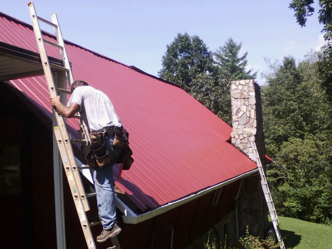 Roofer on a ladder beside a red metal roof and stone chimney — Soffit & Fascia Repair by Roofing Brothers in Louisa